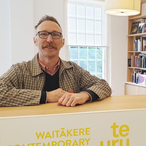 A person wearing glasses, standing behind a counter that says Te Uru Waitākere Contemporary Gallery